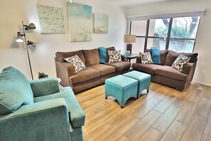 A living room with a brown sectional sofa, teal armchairs, and ottomans, a floor lamp, and artwork on the walls.