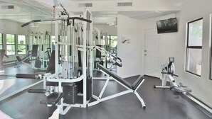 Un gimnasio con diversos equipos de ejercicio, un televisor en la pared y amplios ventanales.