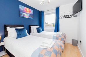 Ein Hotelzimmer mit zwei Betten, einem Gemälde an der Wand, einem an der Wand befestigten Fernseher und einem Fenster mit Vorhängen.