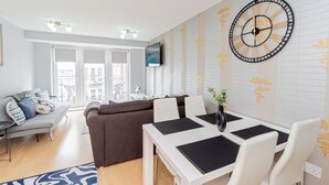 A modern living room with a sofa, a dining table with chairs, a large wall clock, and a TV mounted on the wall. - Modern 2-Bedroom Apartment in Central Basingstoke  walk to Festival Place & The Avil Theatre . (Basingstoke)