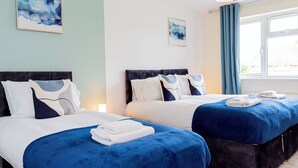 A hotel room with two beds, each with a blue bedspread and white linens. There are two framed abstract paintings on the walls, a window with blue curtains, and a hanging lamp. - 4-Bed Haven in Fawley – Ideal for extended stays, work trips, or family getaways. Spacious, fully furnished, and well-equipped for comfort and convenience. Your perfect home away from home. (Totton)