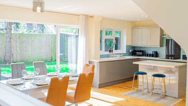 A modern kitchen with a large island, bar stools, and a dining area with a table set for two. - 4-Bed Haven in Fawley – Ideal for extended stays, work trips, or family getaways. Spacious, fully furnished, and well-equipped for comfort and convenience. Your perfect home away from home. (Totton)