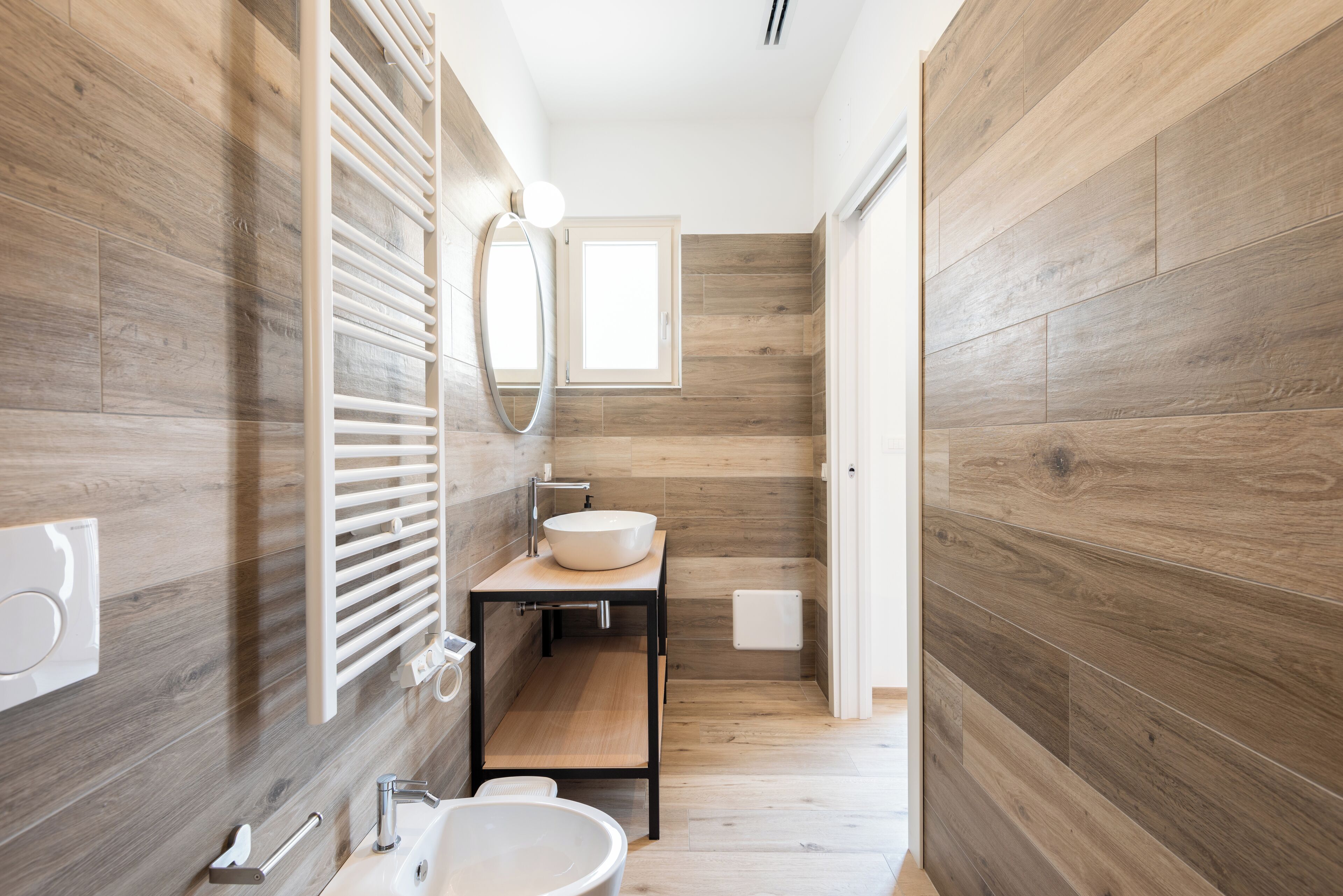 A modern bathroom with wooden flooring, a white sink, a mirror, and a window.