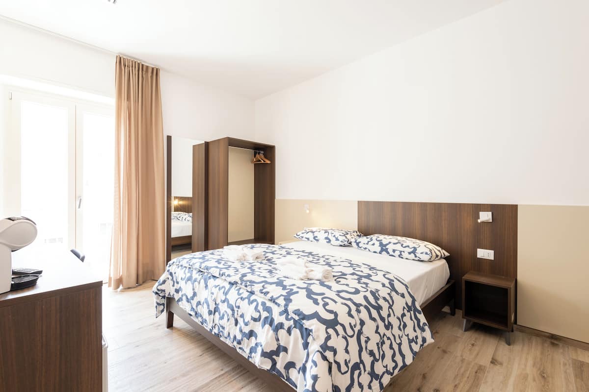 Ein modernes Hotelzimmer mit einem großen Bett, einem Schreibtisch mit Lampe, einem Kleiderschrank und einem Nachttisch.