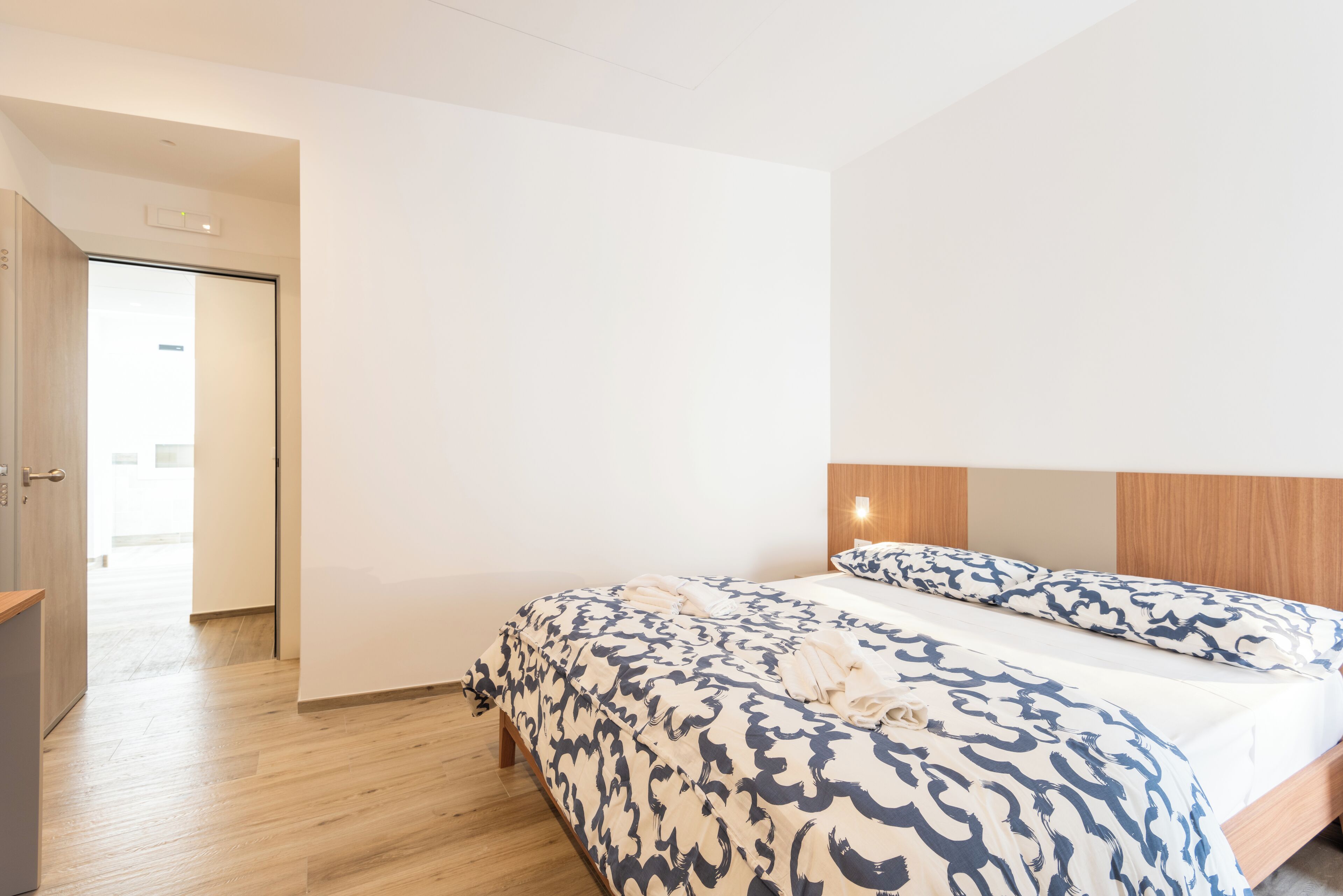 A modern hotel room with a large bed, wooden headboard, and a door leading to a hallway.
