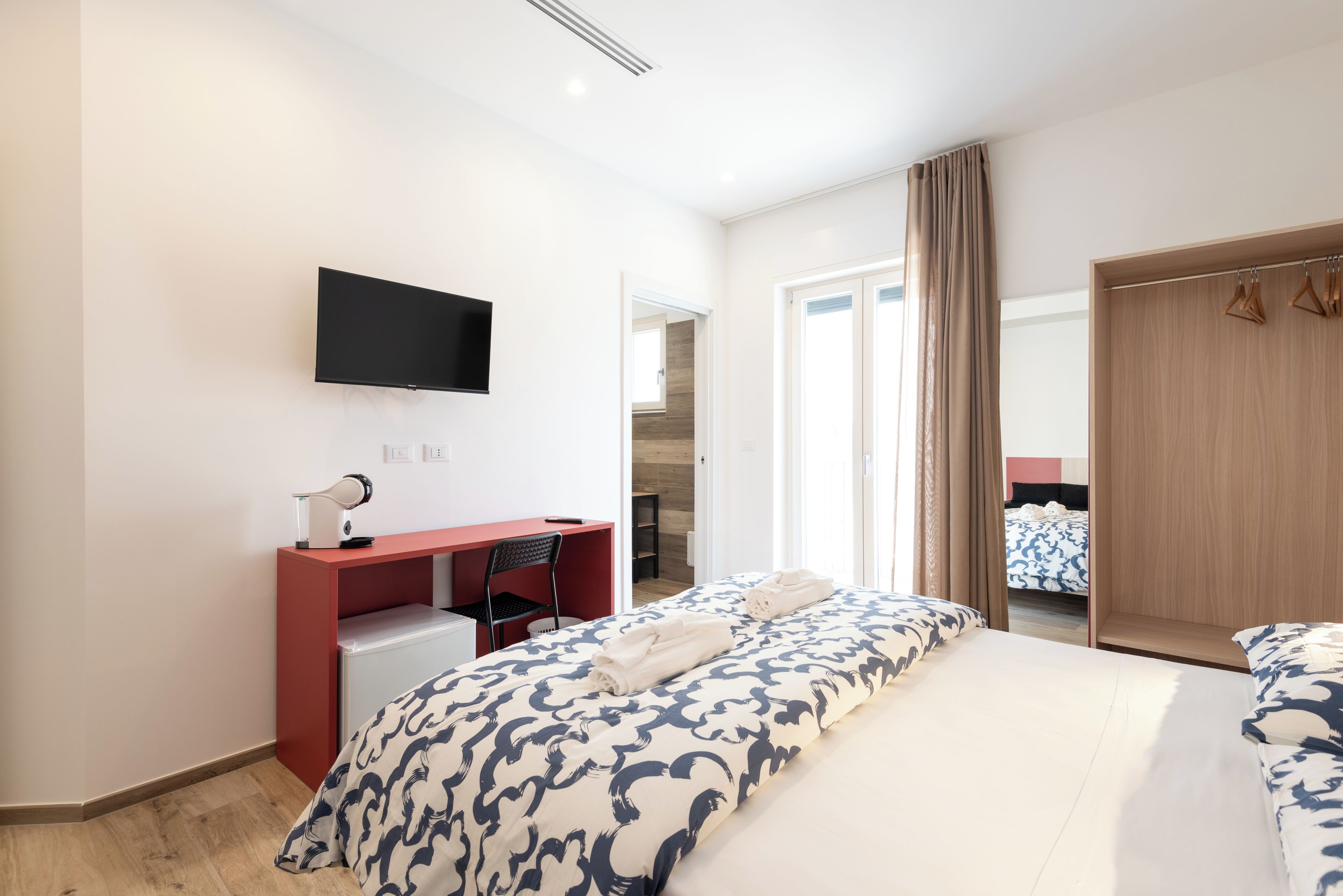 A modern hotel room with a large bed, a desk with a chair, a flat-screen TV, and a wardrobe.
