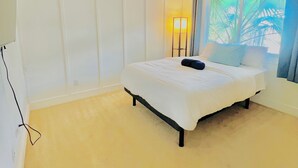 Ein Schlafzimmer mit einem Bett, einer Nachttischlampe, einem Fenster mit Vorhängen und Blick auf Palmen.