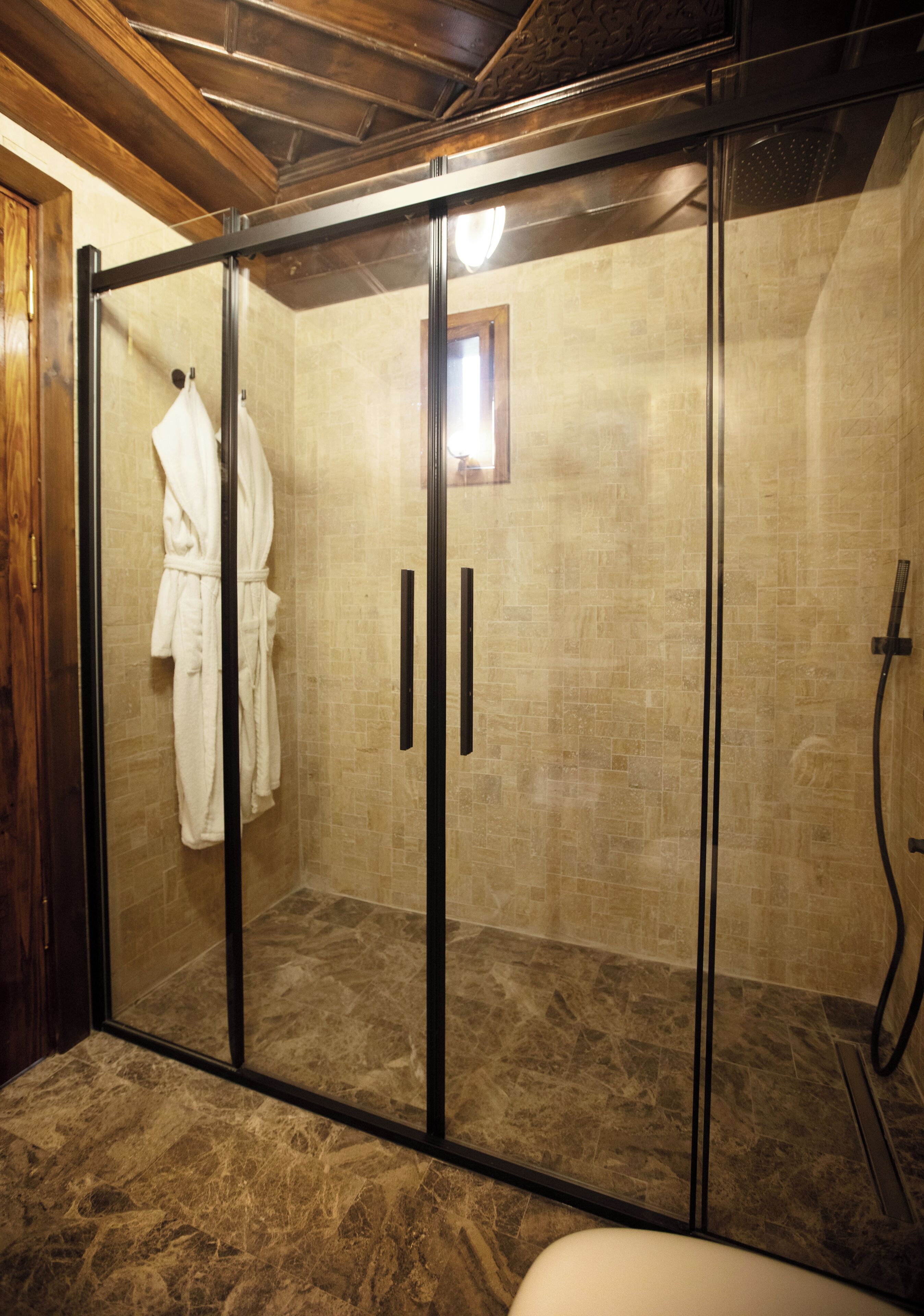 A glass-enclosed shower area with a marble floor, a wooden ceiling, and a white bathrobe hanging on the door.