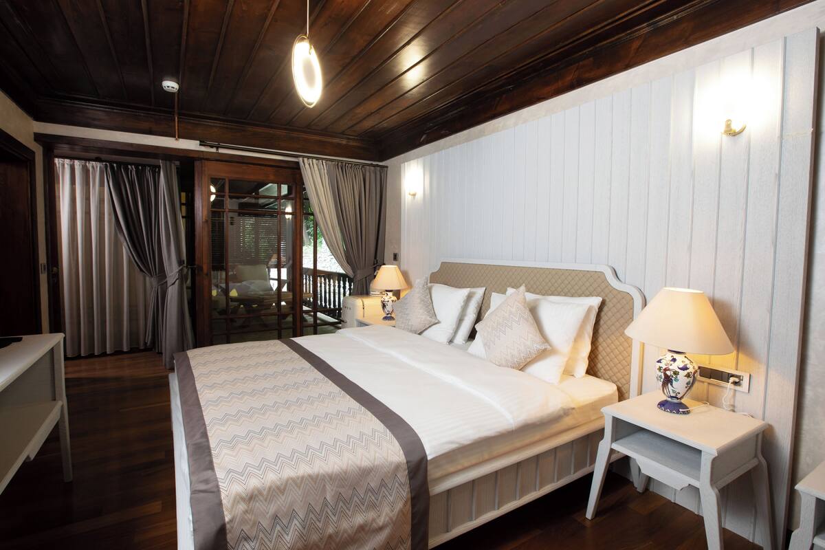 A bedroom with a large bed, two bedside tables with lamps, wooden flooring, and a view through a glass door to an outdoor area.
