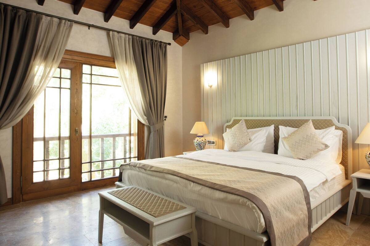 A bedroom with a wooden ceiling, a large bed with white bedding, a nightstand with a lamp, and a window with curtains.
