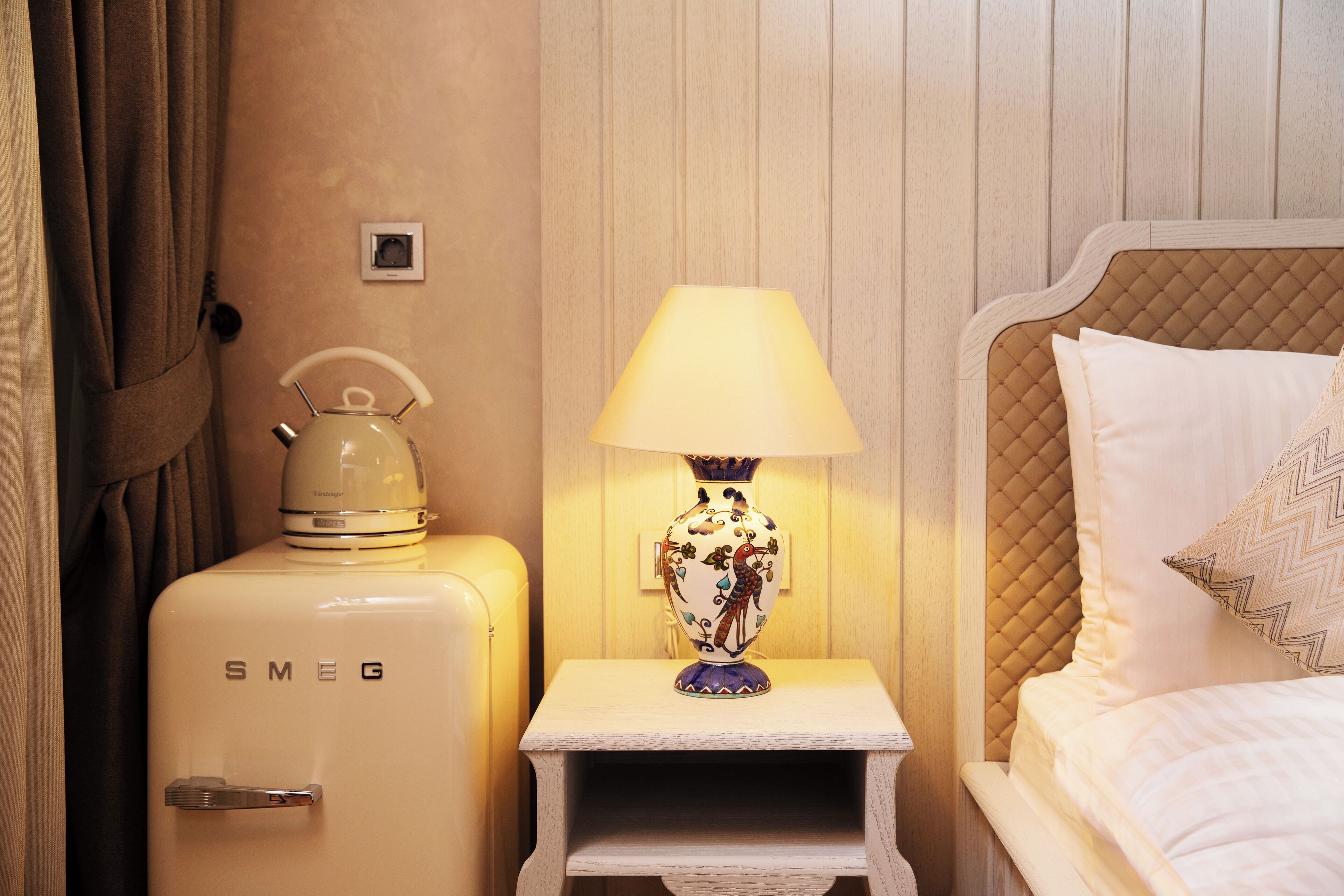 A hotel room with a vintage Smeg refrigerator, a bedside lamp, and a bed with a quilted headboard.
