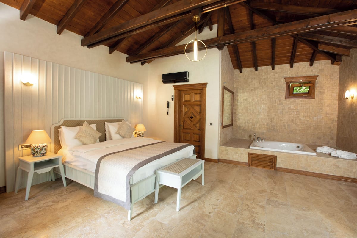 A bedroom with a bed, bedside tables, a bench, a wooden door, a wall-mounted TV, and a bathroom with a bathtub and a shower.