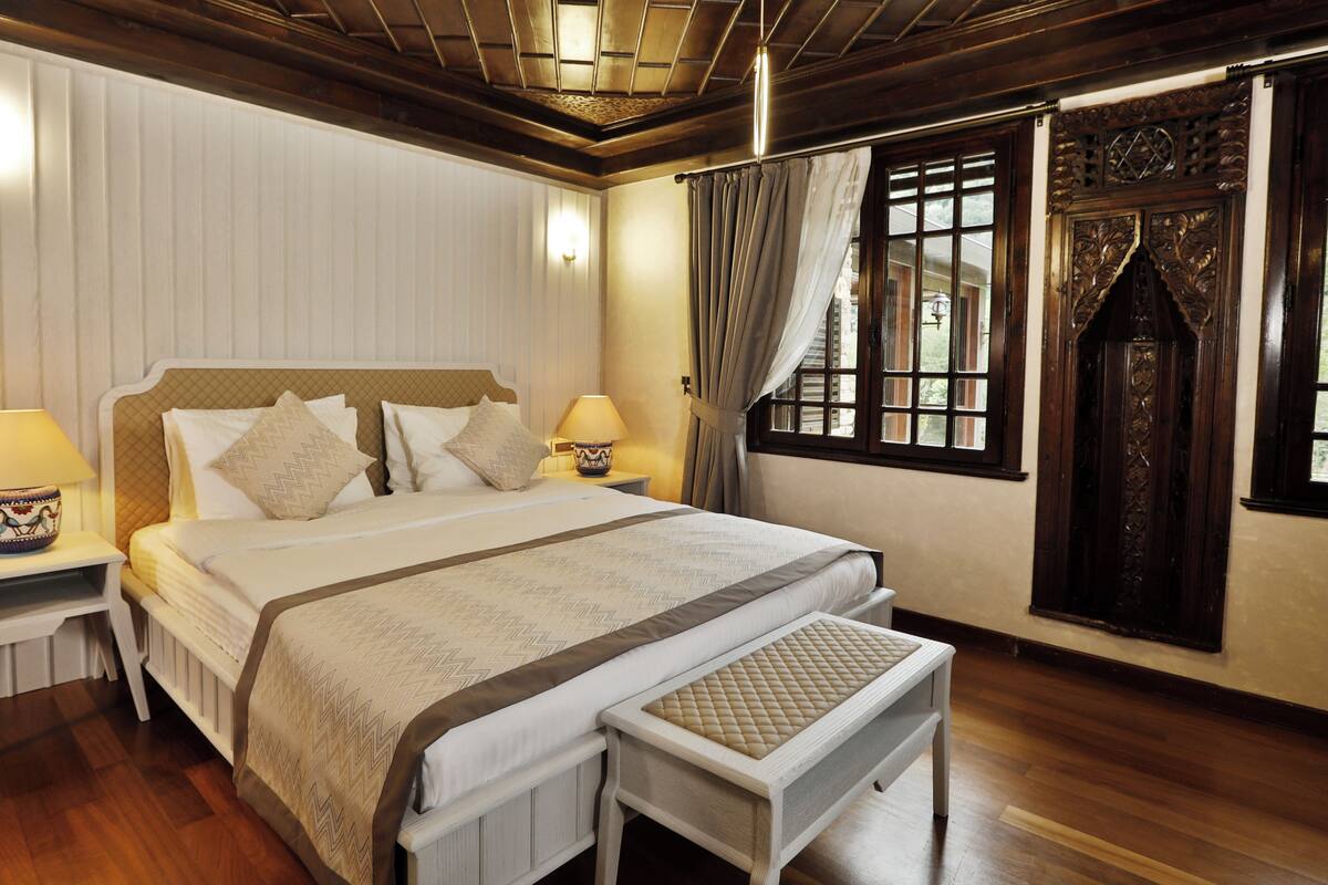 A neatly arranged bedroom with a large bed, two bedside tables with lamps, a bench at the foot of the bed, and a window with curtains.