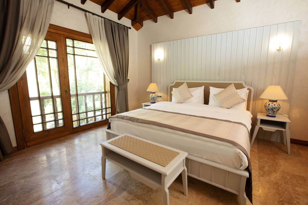 A bedroom with a large bed, two bedside tables with lamps, a bench, and a view through a glass door.