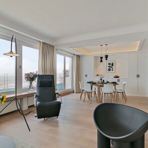 A modern living room with a black chair, a dining table with chairs, and a view of the sea through large windows. - Panoramic Seaview Apartment in t Zoute (Knokke-Heist)