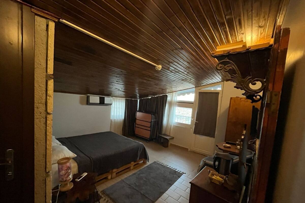 Un dormitori amb sostre de fusta, un llit, un calaixera, un mirall i una finestra amb cortines.