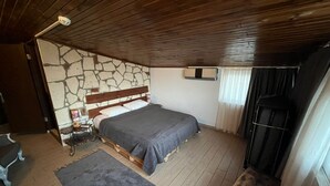 A bedroom with a stone wall, wooden ceiling, a bed with a grey bedspread, a nightstand, a lamp, a window with curtains, and a wooden bench. - hotel budapest (Seferihisar)