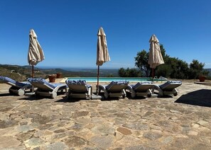 A stone patio with lounge chairs and umbrellas overlooking a pool and a scenic view.