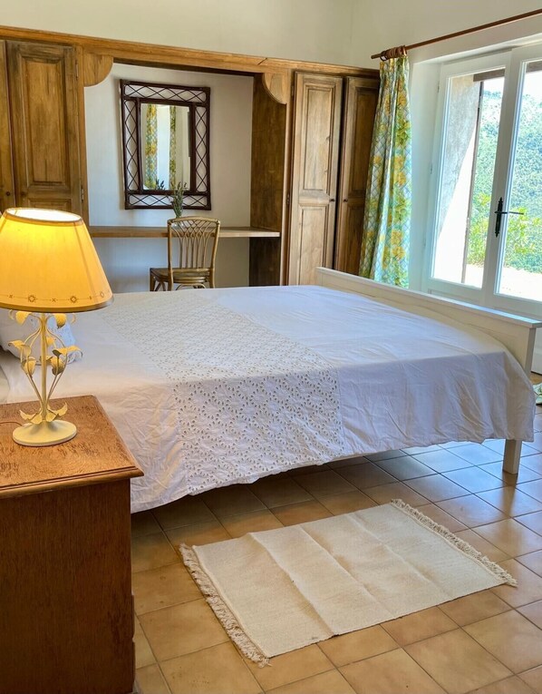 A bedroom with a large bed, a bedside table with a lamp, a chair, a mirror, and a window with curtains.