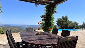 Une table en bois, entourée de chaises, placée sous une pergola, avec vue sur une piscine et des montagnes.
