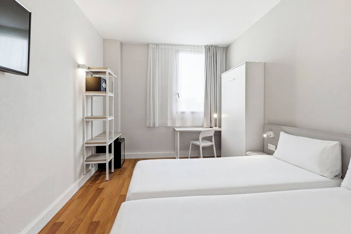 Una camera d'albergo con due letti, una scrivania, una sedia, un armadio e una finestra con tende.