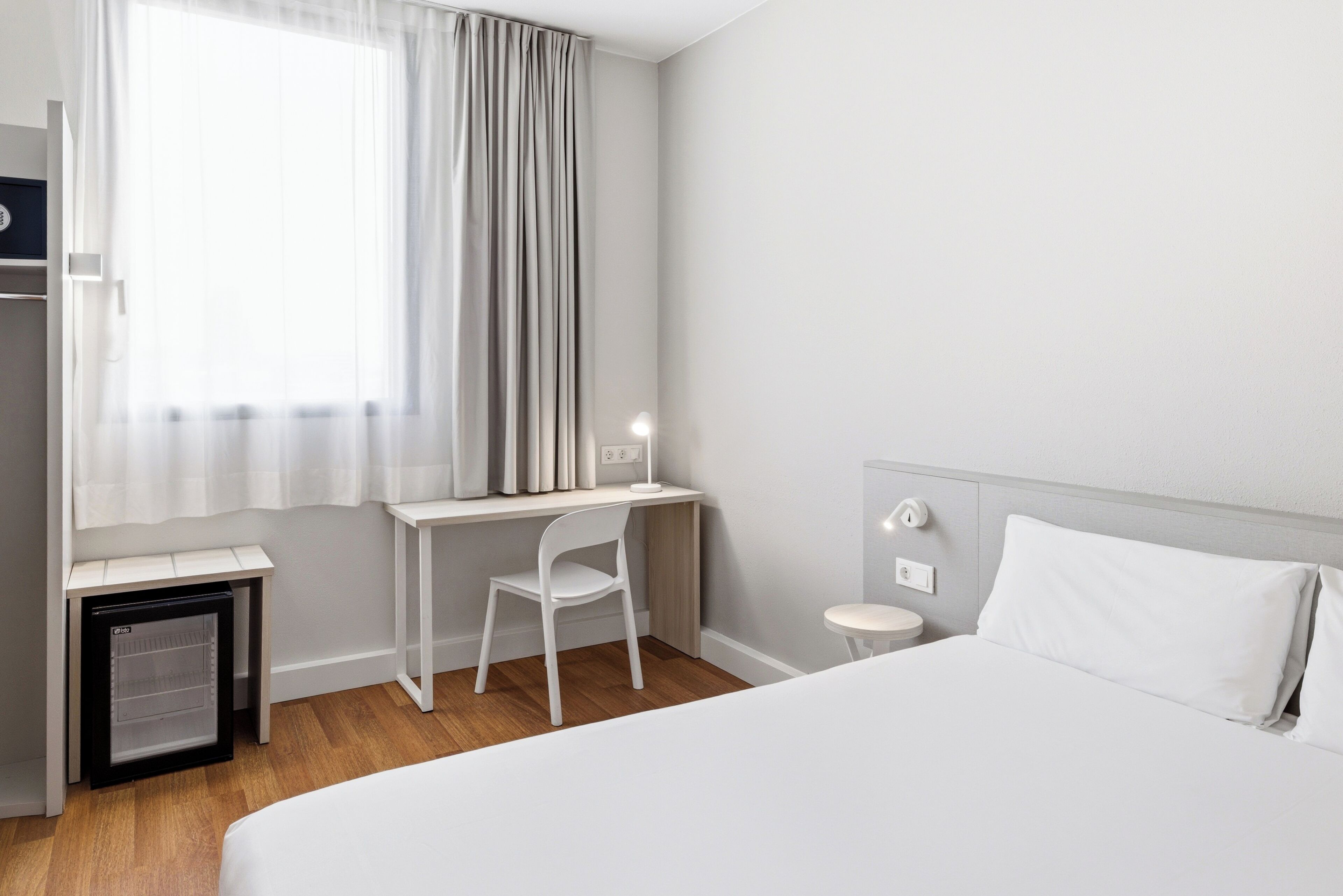 A hotel room with a bed, a desk with a chair, a small refrigerator, and a window with curtains.