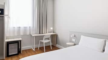 A hotel room with a bed, a desk with a chair, a small refrigerator, and a window with curtains.