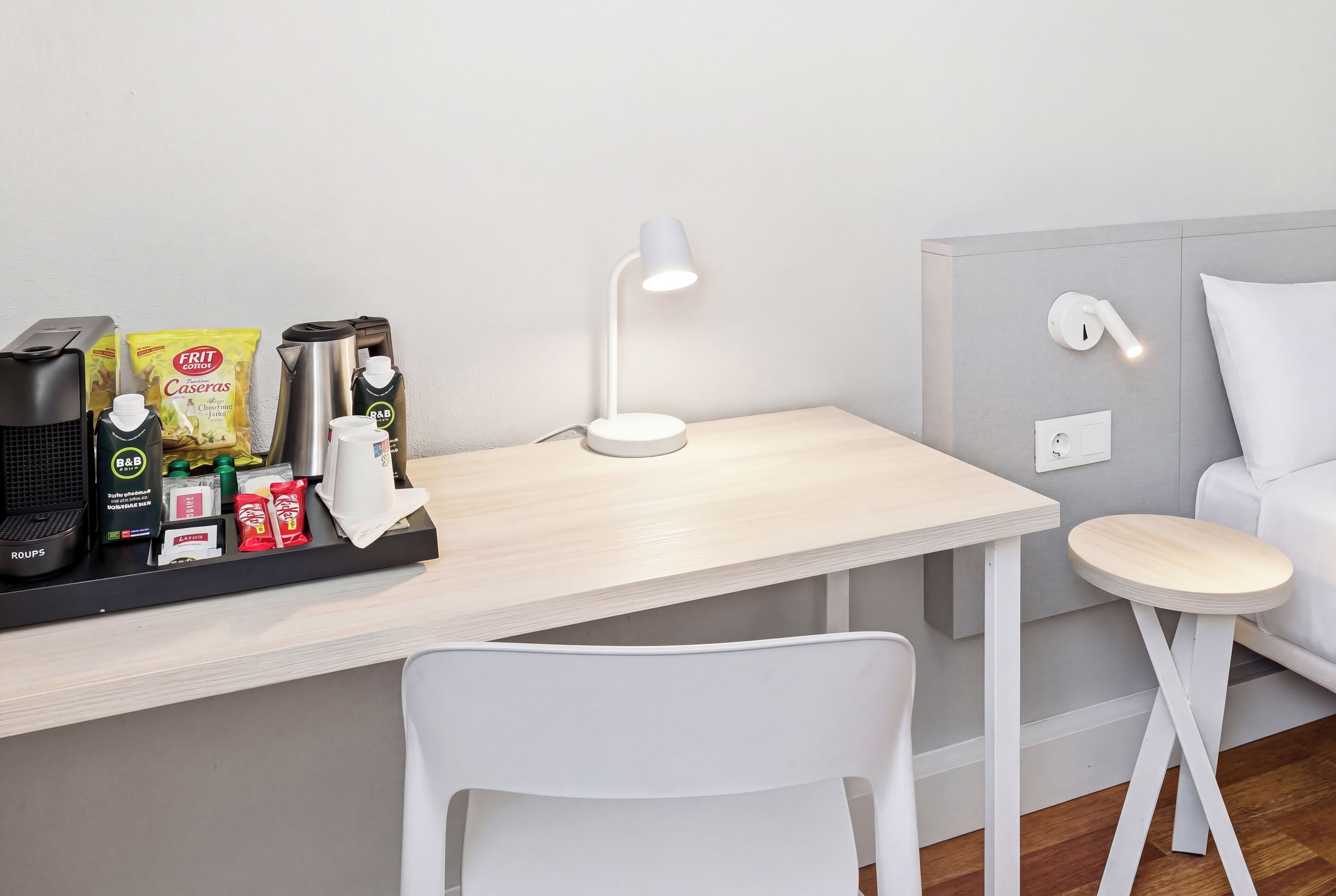 Een kleine, moderne kamer met een bureau, een witte stoel, een koffiezetapparaat, een waterkoker en een lamp.