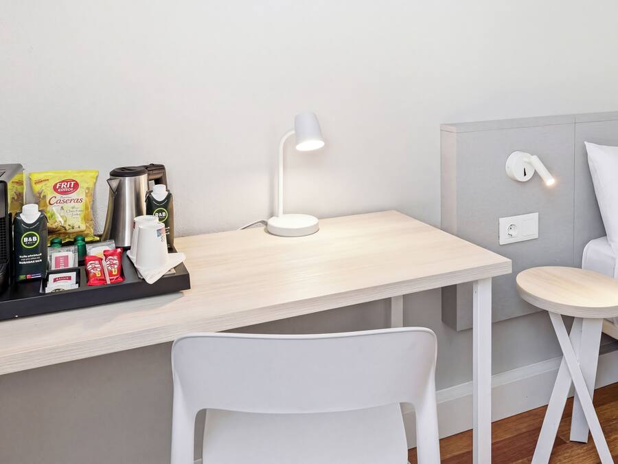 A small, modern room with a desk, a white chair, a coffee maker, a kettle, and a lamp.