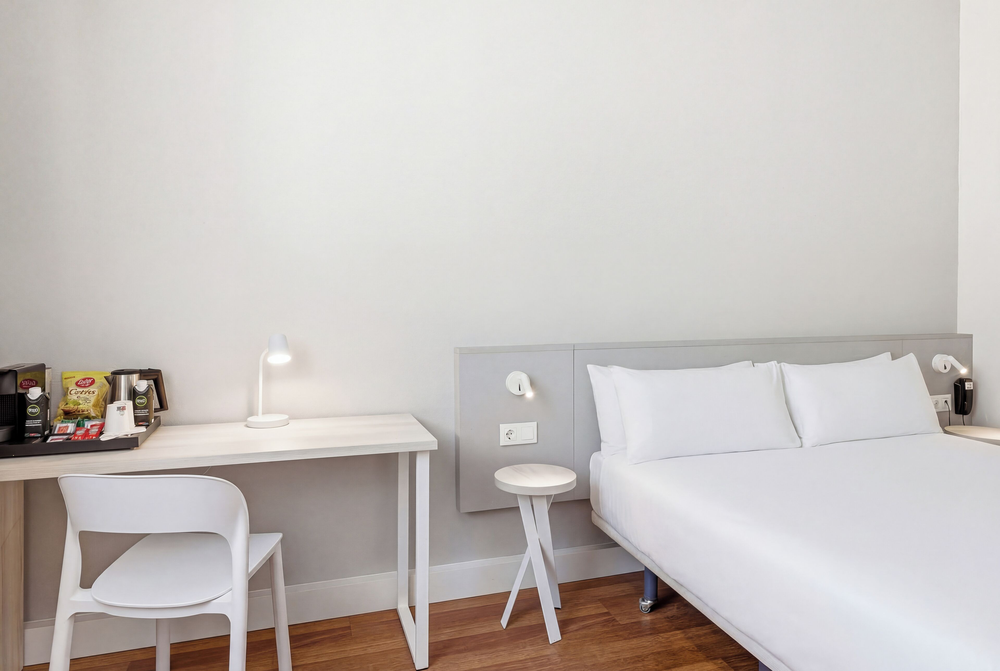 A hotel room with a bed, a desk, a chair, a small table, and a wall-mounted light.