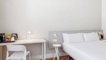 A hotel room with a bed, a desk, a chair, a small table, and a wall-mounted light.