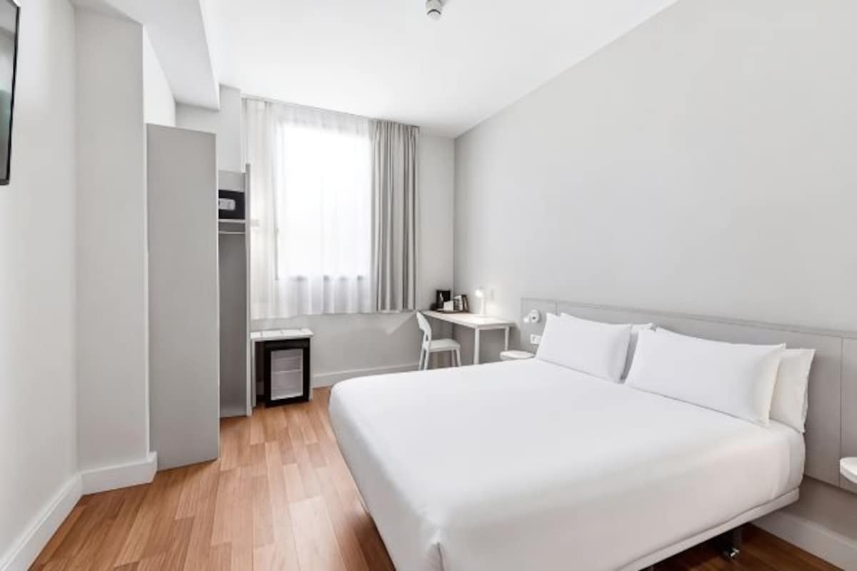 Ett hotellrum med en säng, ett skrivbord, en stol och ett fönster med gardiner.