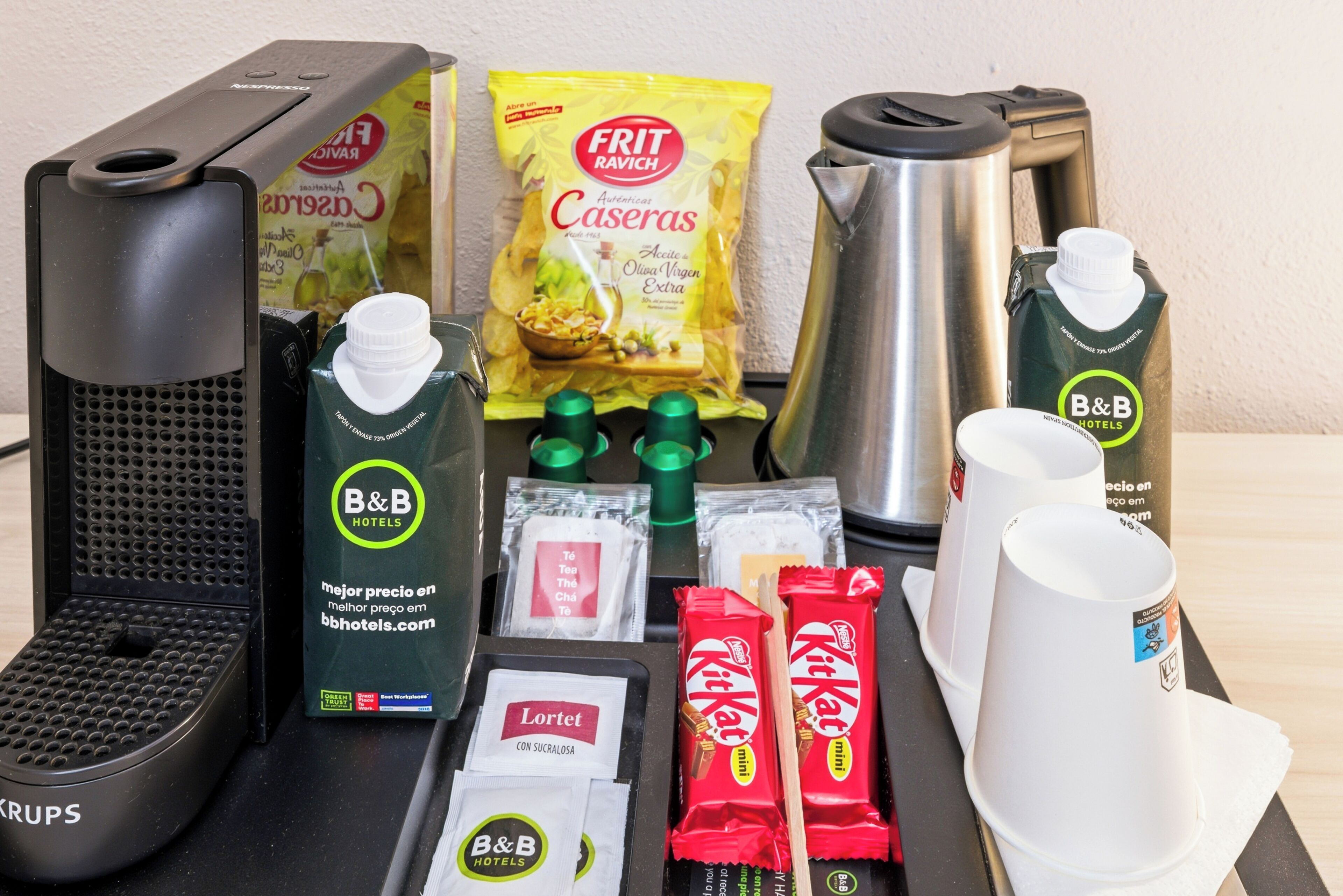 Ontbijtbuffet met een koffiezetapparaat, een fles B&B Hotels sinaasappelsap, een zakje Frit Ravich caseras chips, een zakje Loretet met sucralose, twee KitKat chocoladerepen en een thermosfles.