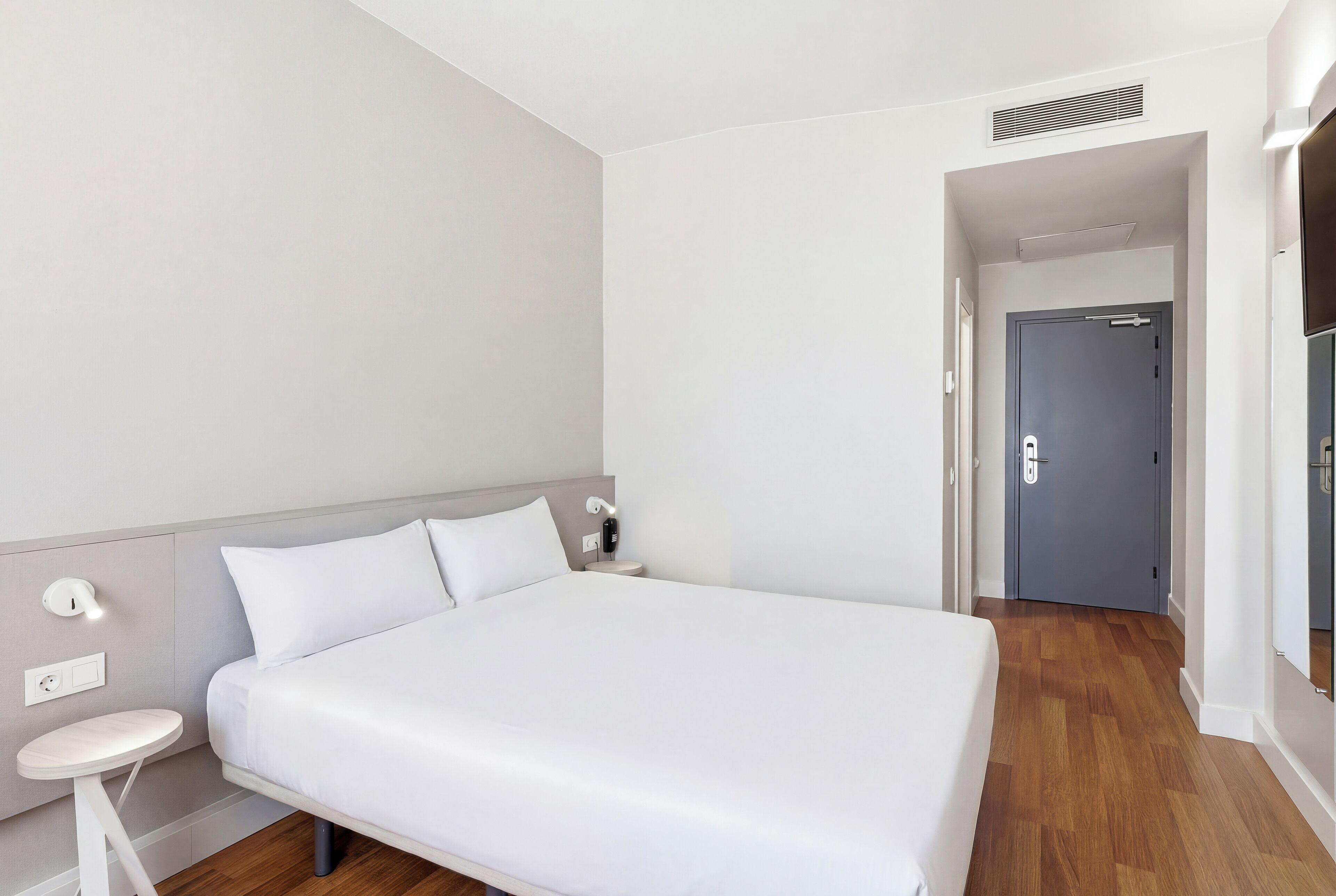 A modern hotel room with a large bed, bedside table, and a door leading to another room.