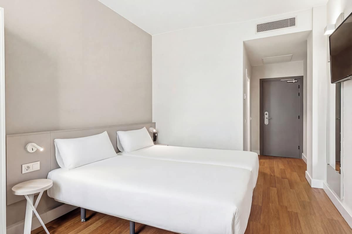 Una camera d'albergo moderna con un letto matrimoniale, comodini, una TV e una porta che dà su un corridoio.