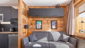 A modern living room with a grey sofa, wooden walls, a kitchen area with a dishwasher, and framed pictures on the wall.