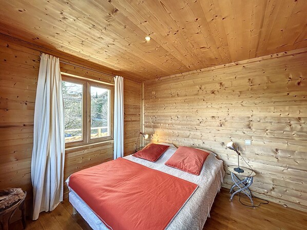 Een slaapkamer in een houten chalet, met een bed, nachtkastje, lamp en raam met gordijnen.