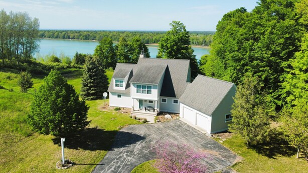 A two-story house with a steeply pitched roof, surrounded by lush greenery and a lake.