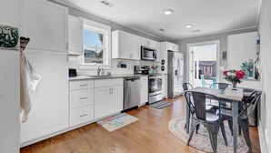 A modern kitchen with white cabinetry, stainless steel appliances, and a dining area with a table and chairs. - Turtle Time- Charming 3BR Getaway Near Kure Beach (Kure Beach)