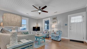 A living room with a ceiling fan, a television, a sofa, a coffee table, and a door. - Turtle Time- Charming 3BR Getaway Near Kure Beach (Kure Beach)