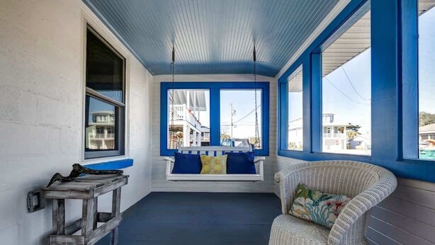 A porch with a swing, a wicker chair, and a wooden table. - Turtle Time- Charming 3BR Getaway Near Kure Beach (Kure Beach)