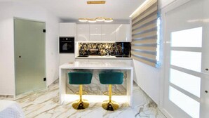 Cocina moderna con armarios blancos, encimera de mármol y horno empotrado. Hay dos taburetes verdes con patas doradas.