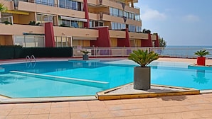 A swimming pool with a tiled deck, surrounded by a building with balconies and a view of the sea.