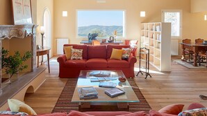 A spacious living room with a red sofa set, a glass coffee table, and a fireplace. There is a bookshelf and a dining area visible in the background.
