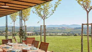An outdoor dining area with a table set for six, surrounded by wooden chairs, under a wooden pergola with a view of rolling hills and trees.