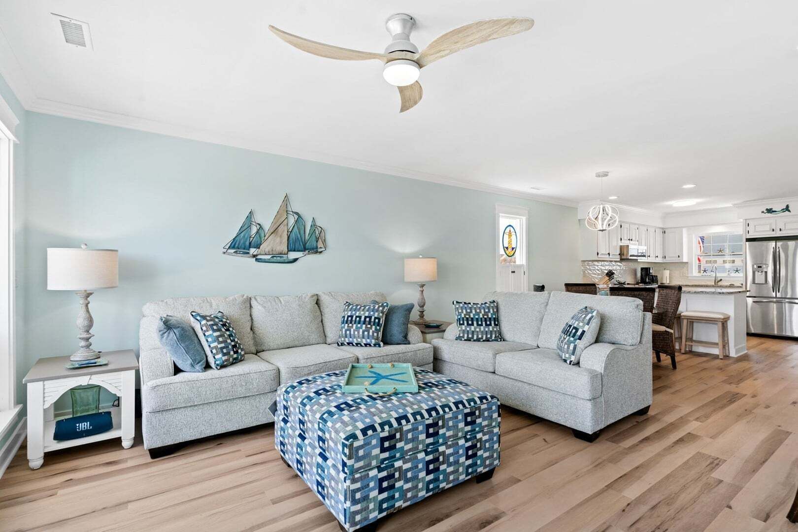 Ein Wohnzimmer mit Ecksofa, einem gemusterten Ottomanen, Deckenventilator und maritimen Wandbildern.