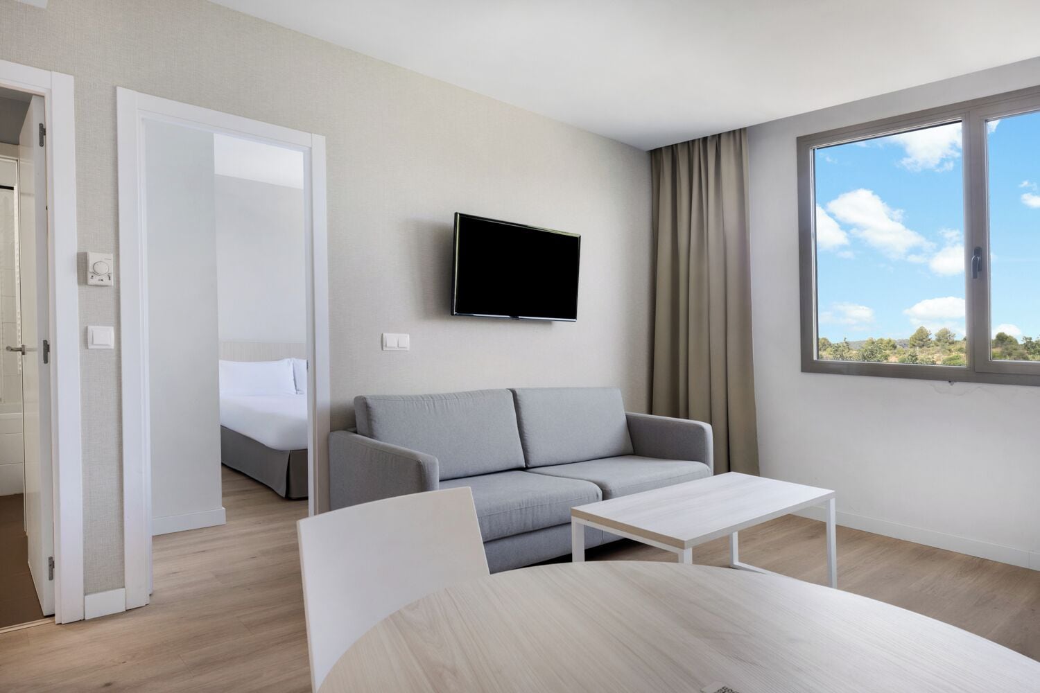A modern hotel room with a grey sofa, a flat-screen TV, a small white table, and a large window with a view of greenery.
