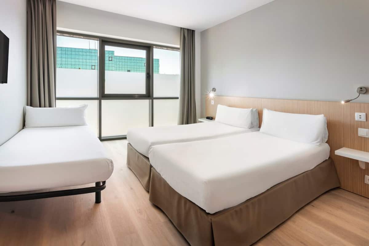 Habitación de hotel con dos camas, una mesita de noche, una lámpara y una ventana con vistas a un edificio.