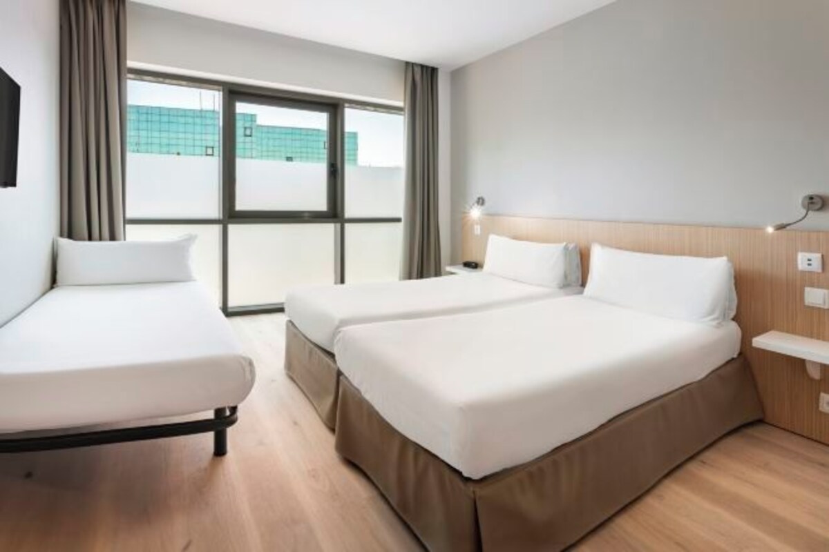 Habitación de hotel con dos camas, una ventana con cortinas, una mesita de noche con lámpara y vista a un edificio.