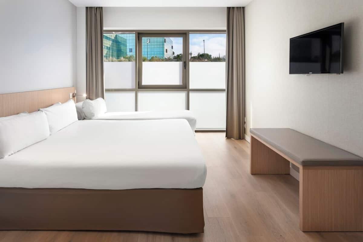 Habitación de hotel con dos camas, un banco, un televisor y ventanas amplias con vista a edificios.